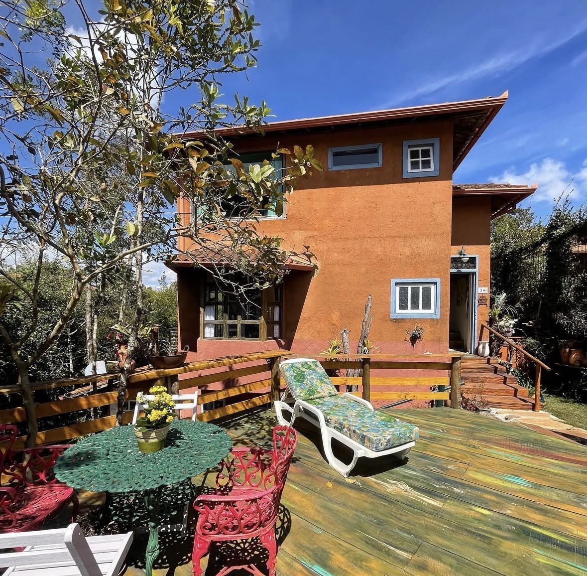 Fachada da Casa das Orquídeas com deck colorido, cadeiras e espreguiçadeira sob árvore